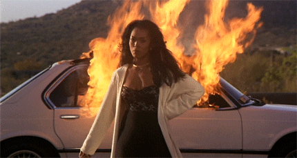 GIF of a woman walking away from a car on fire and pumping her fist